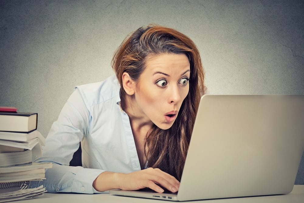 Portrait young shocked business woman sitting in front of laptop computer looking at screen isolated grey wall background. Funny face expression emotion feelings problem perception reaction 